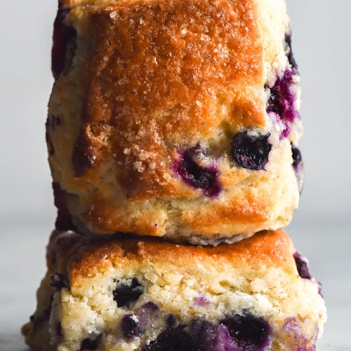 An side on view of two gluten free blueberry scones stacked on top of each other on a white table against a white background. The bottom scone sits with it's base against the table, revealing the fluffy crumb and oozy blueberries inside. The second scone sits with it's front towards the camera, showing off it's golden top speckled with finishing sugar and blueberries