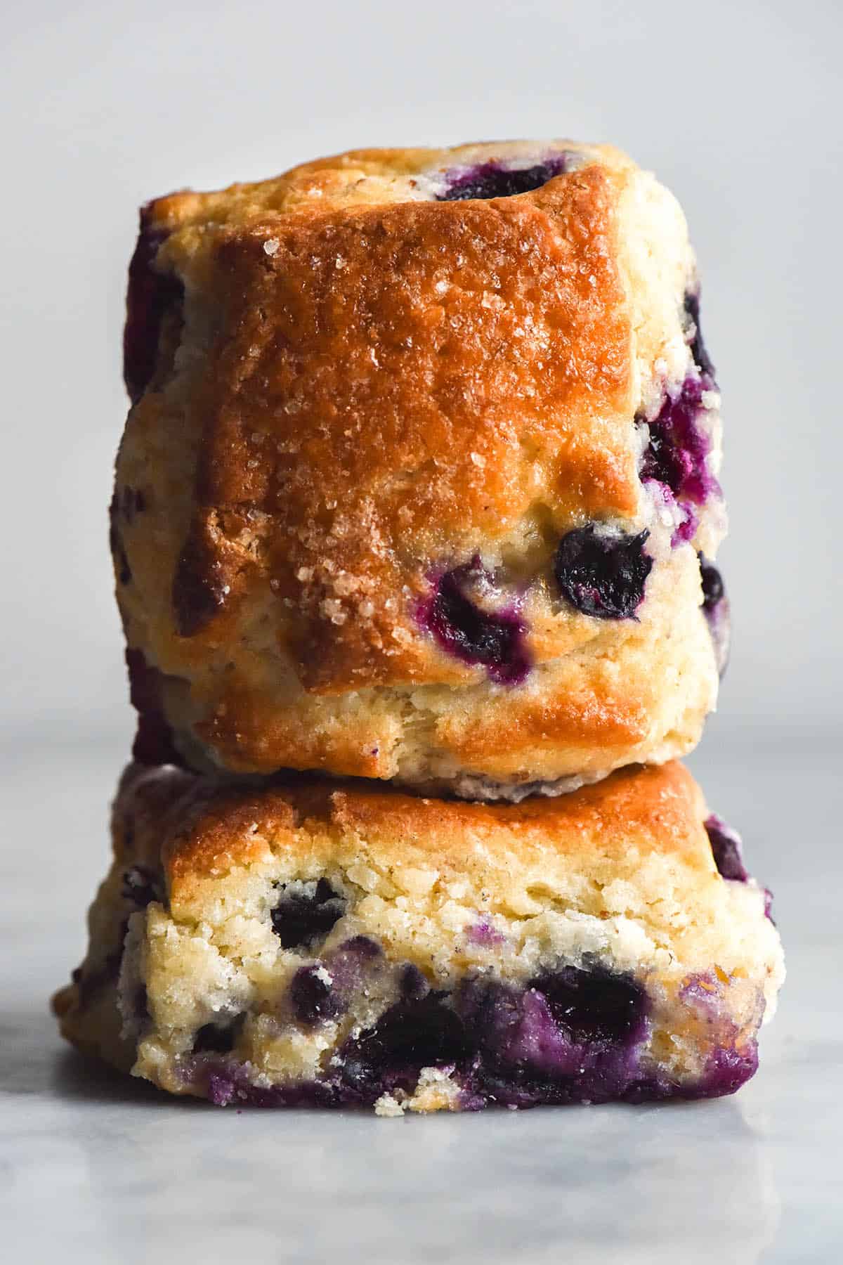 An side on view of two gluten free blueberry scones stacked on top of each other on a white table against a white background. The bottom scone sits with it's base against the table, revealing the fluffy crumb and oozy blueberries inside. The second scone sits with it's front towards the camera, showing off it's golden top speckled with finishing sugar and blueberries