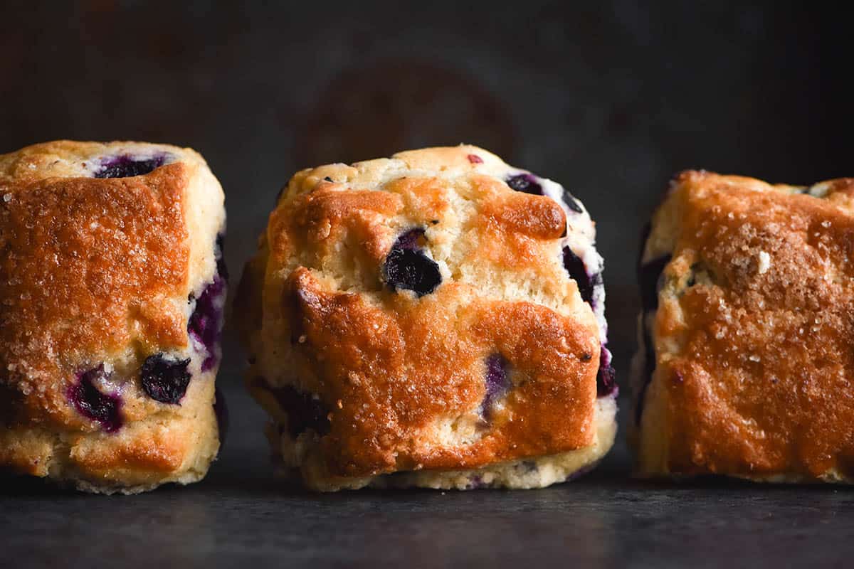 A side on close up photo of three gluten free scones propped up so their tops are facing the camera. They are square scones that sit evenly spaced apart on a dark metal table against a dark metal backdrop. The scone tops are golden brown and speckled with finishing sugar and the oozy purple colour of cooked blueberries