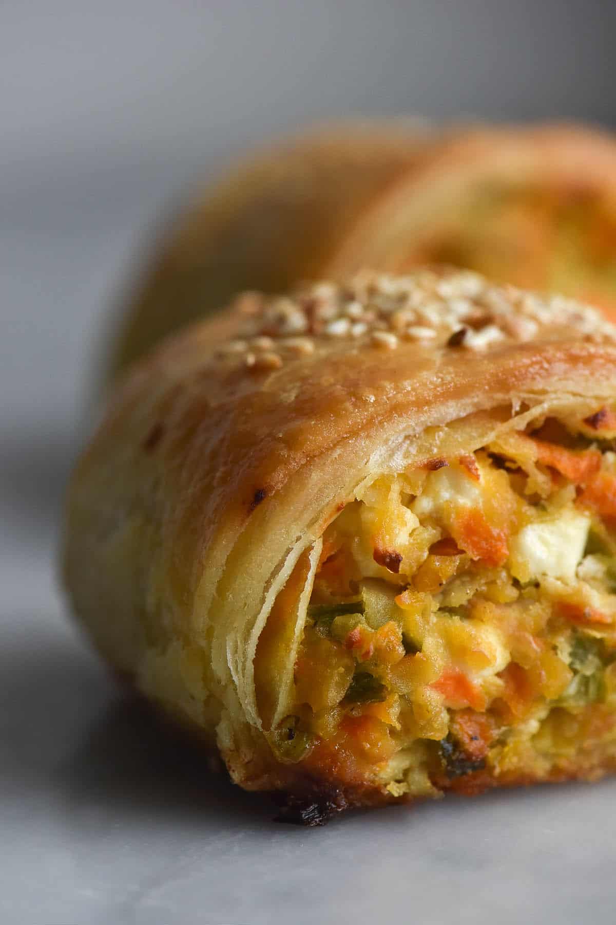 A very close up aerial view of a gluten free chickpea, vegetable and feta sausage roll. The roll sits atop a white marble table with a white backdrop, and a second roll sits in the background. The photo focuses on the flaky and layered pastry that encapsulates the vegetable filling. A bit of feta peeks through the vegetables and sesame seeds nestle on top of the roll