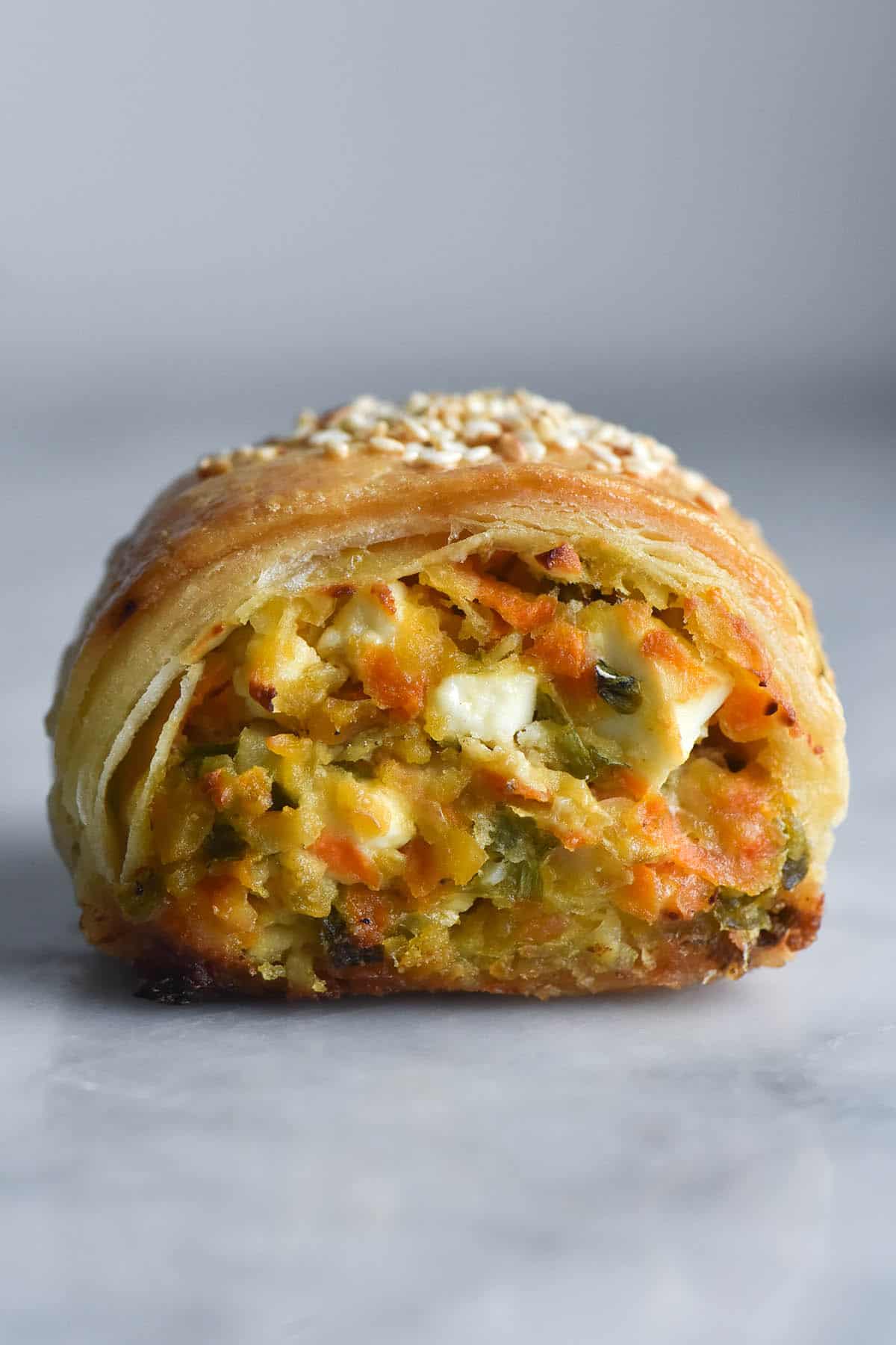 A side on close up view of a gluten free chickpea, vegetable and feta sausage roll. The roll sits atop a white marble table against a white backdrop. The side on view exposes the cooked mixed vegetable innards with chunks of feta peeking through. On top, the pastry is flaky and layered, dotted with some toasted sesame seeds which are visible