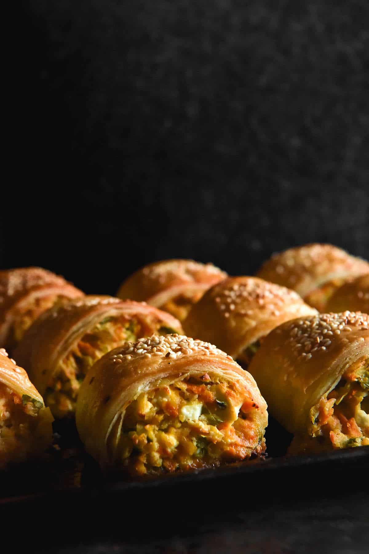 A side on, moody image of rows of gluten free chickpea, vegetable and feta sausage rolls. The rolls sit on a dark baking tray against the torso of a person wearing a dark grey fluffy jumper in the background. From the side on angle you can see the vegetables and feta peeking out from underneath flaky pastry topped with sesame seeds