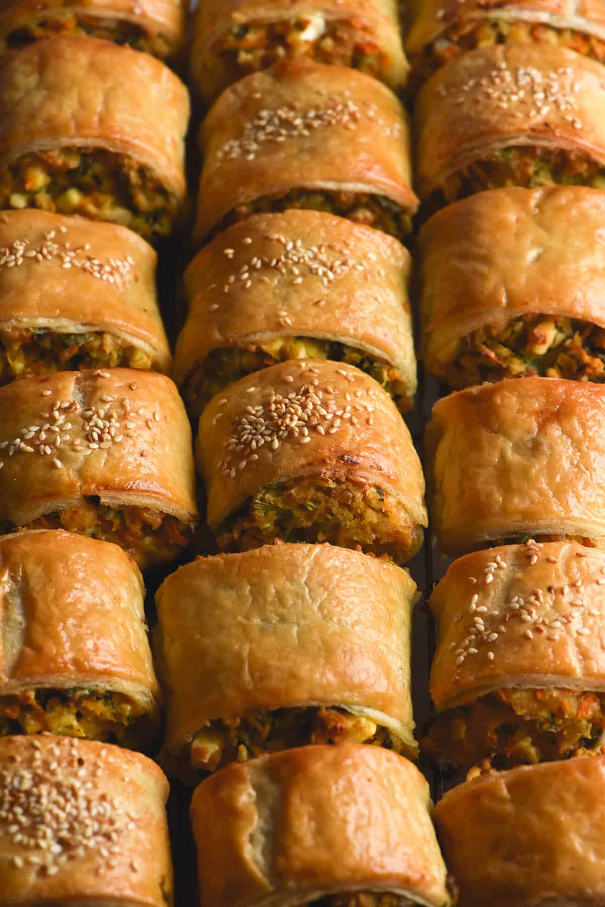 An aerial, close up view of rows of gluten free chickpea, vegetable and feta sausage rolls. The rolls are golden brown and topped with toasted sesame seeds. They hug each other tightly so no background can be seen underneath