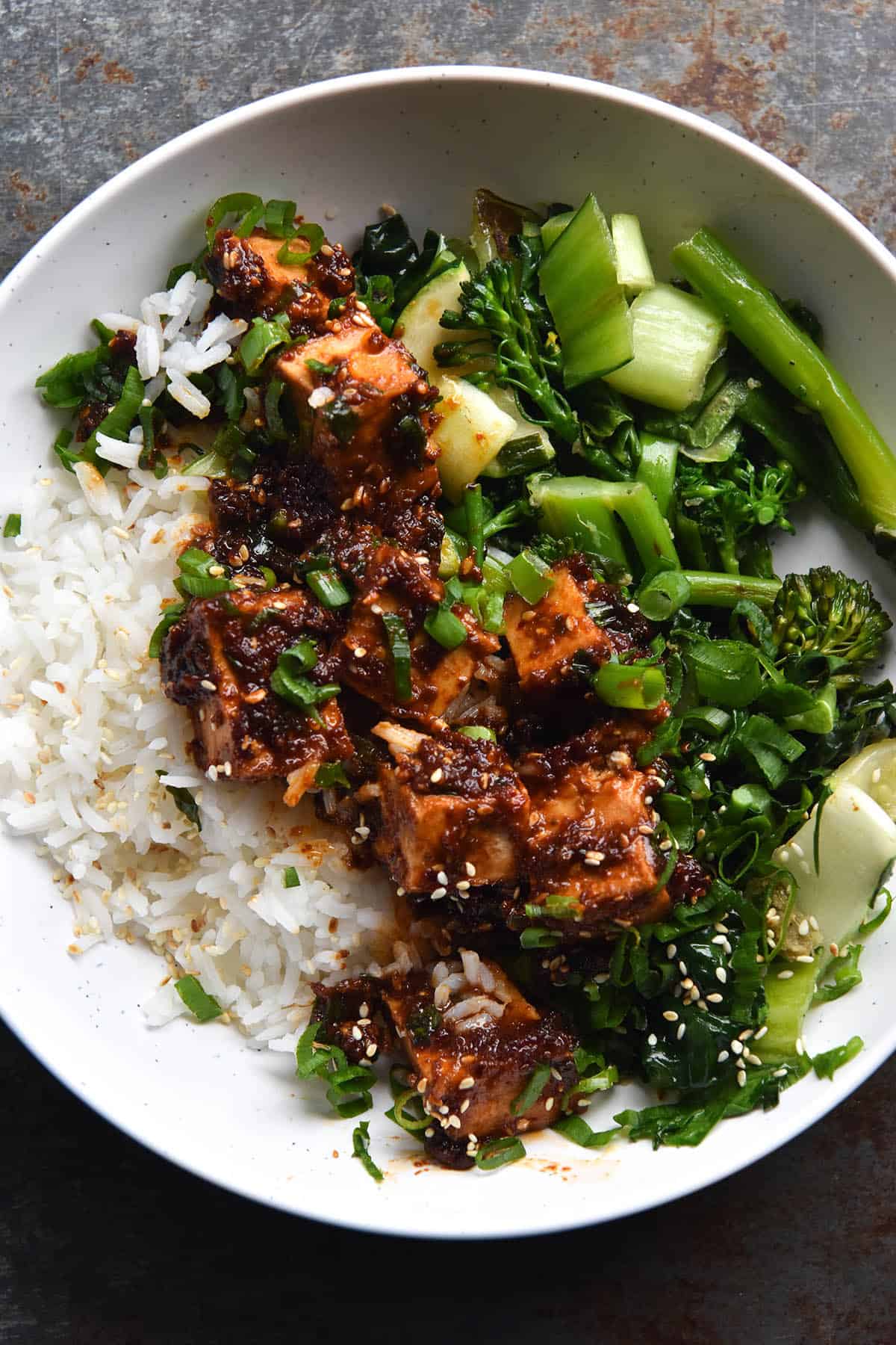 An aerial close up view of a bowl of chilli oil tofu on a bed of asian greens and white rice. The bowl is casually assembled, with greens to the right, tofu in the centre and rice on the left. The bowl is white and sits atop a medium blue metal background