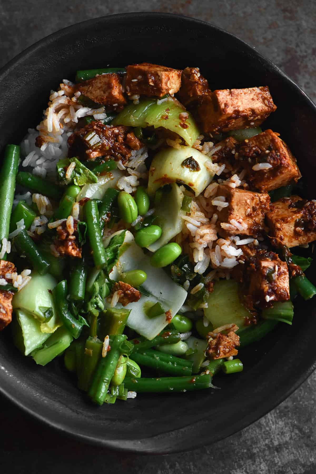 An aerial close up view of a plate of chilli oil tofu, Asian greens and rice that have been mixed together and look casually plated. The meal is served in a dark grey ceramic bowl and sits against a mottled dark silver metal backdrop