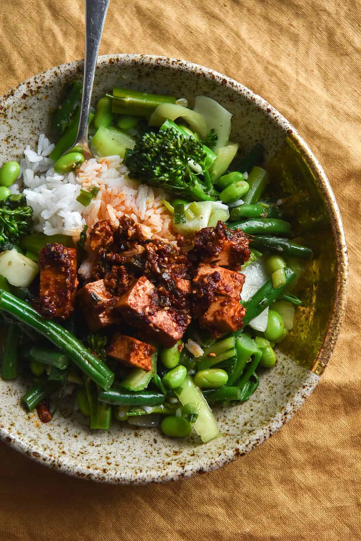 An aerial close up view of a bowl of chilli oil tofu, Asian greens and rice against a rust coloured linen tea towel backdrop. The bowl is a beige ceramic speckled one and the meal is casually arranged inside. A fork sits in the top left of the bowl and extends up beyond the top of the image