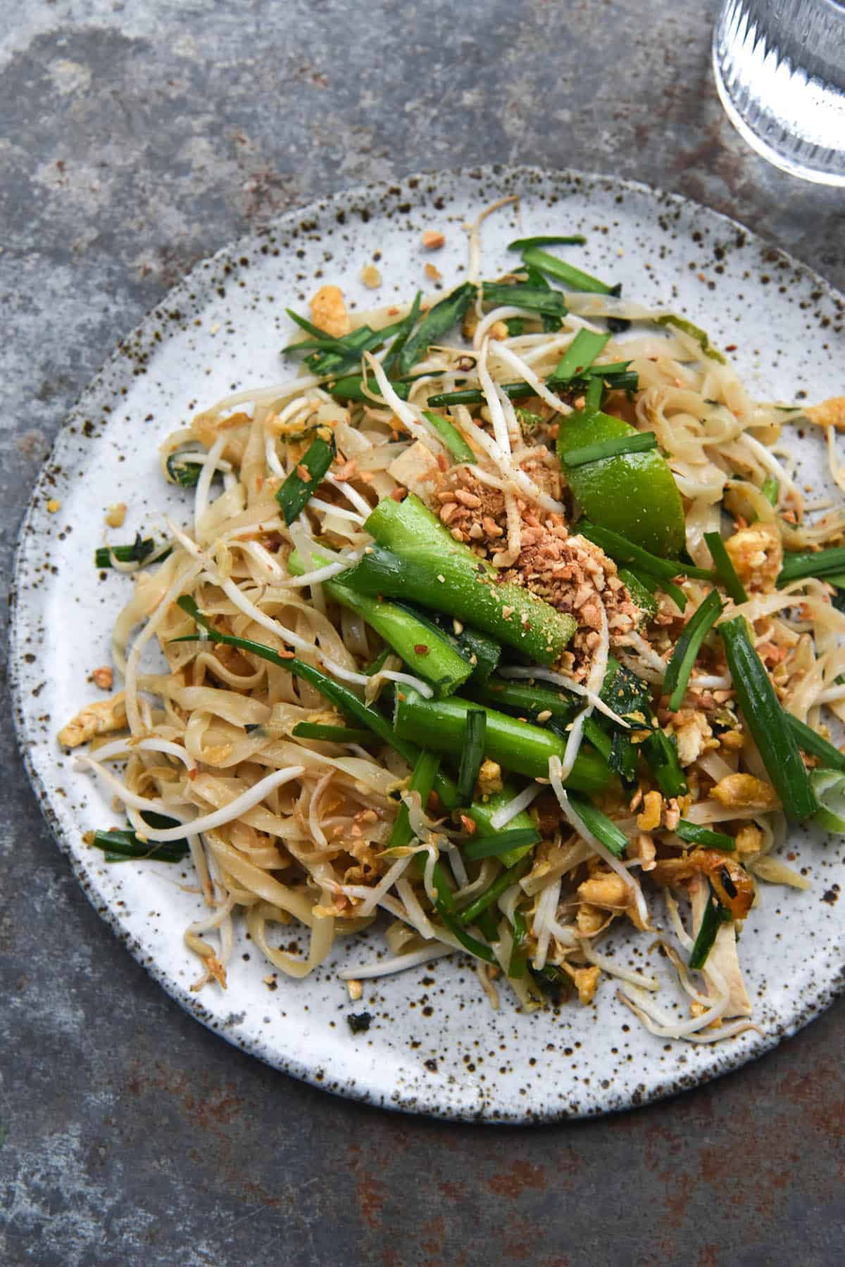 An aerial view of a white speckled plate of FODMAP friendly vegetarian or vegan Pad Thai. The pad Thai is casually arranged on the plate and topped with spring onion greens, toasted peanuts and beansprouts. The plate sits atop a denim blue mottled metal backdrop