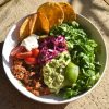 A side on brightly lit view of a plate of FODMAP friendly vegetarian chilli served with salad, guacamole, a wedge of lime, some fodmap friendly 'pickled red onion', corn chips, sour cream and tomatoes. It sits atop terracotta tiles in bright sunlight, with the shadow of some palm leaves in the background of the image