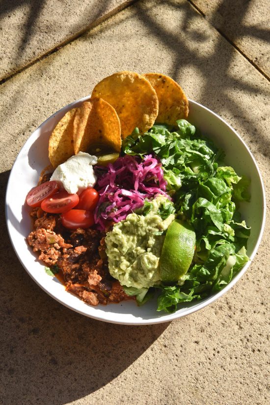 A side on brightly lit view of a plate of FODMAP friendly vegetarian chilli served with salad, guacamole, a wedge of lime, some fodmap friendly 'pickled red onion', corn chips, sour cream and tomatoes. It sits atop terracotta tiles in bright sunlight, with the shadow of some palm leaves in the background of the image
