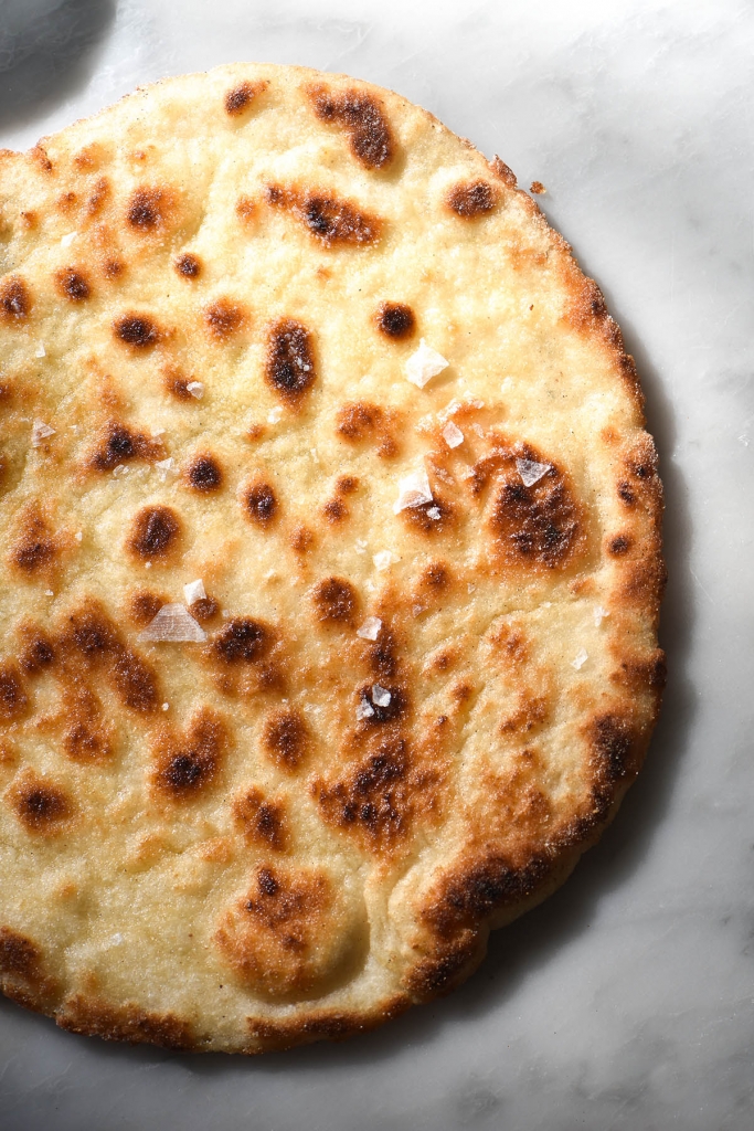 An aerial close up shot of a gluten free yeasted flatbread on a brightly lit marble table. The flatbread is golden brown and darkened in some spots, and has a sprinkle of sea salt flakes on top