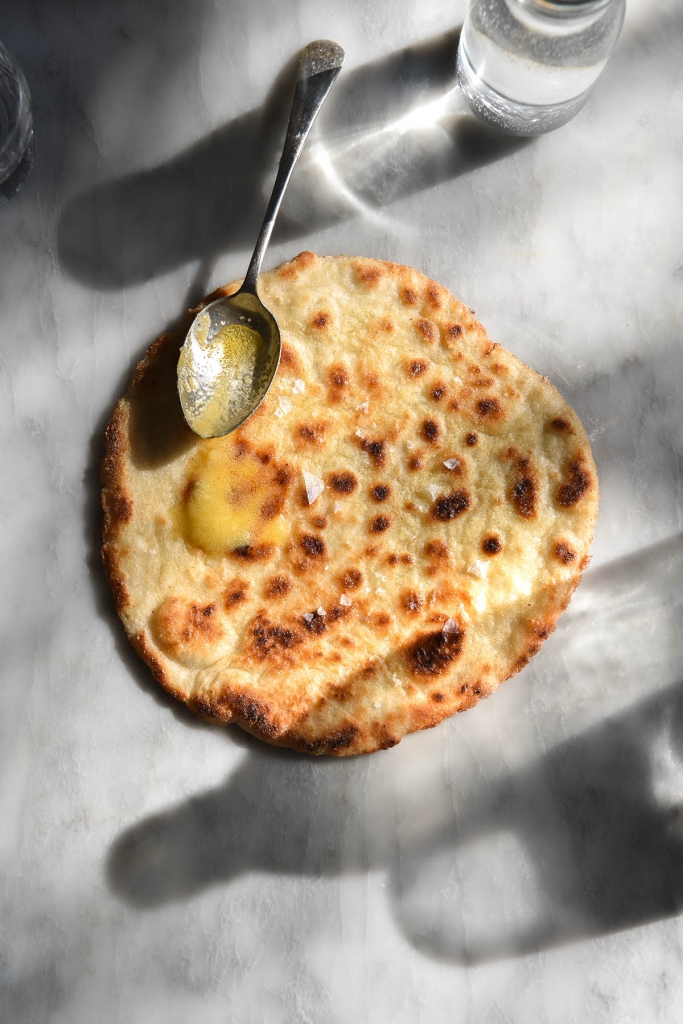 An aerial view of a brightly lit gluten free yeasted flatbread atop a white marble table. The table is scattered with glasses and water bottles which create bright shadows against the marble. A spoon drizzles garlic infused ghee over the top of the flatbread