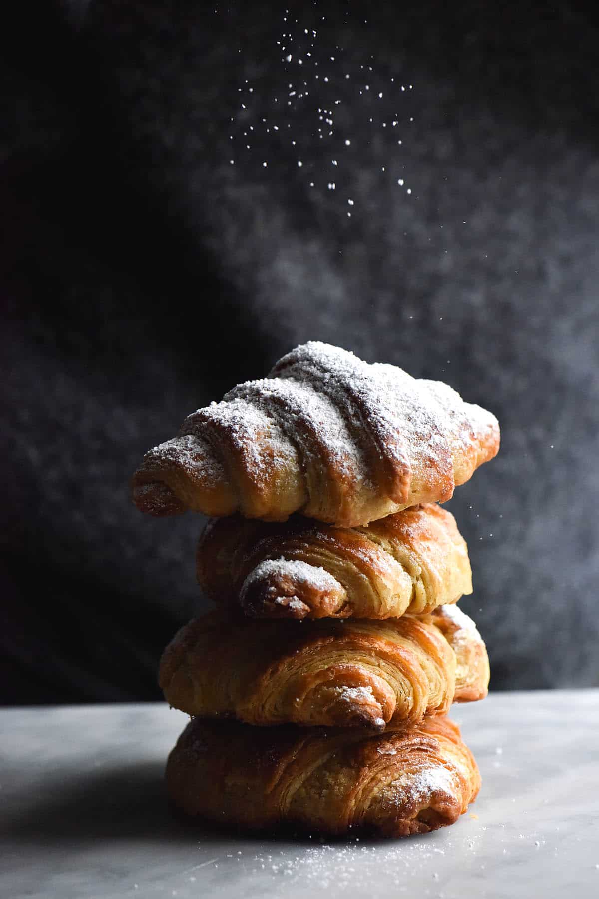 A side on close up view of a stack of gluten free croissants sitting on a white marble table. The background is the torso of a person wearing a dark grey fluffy jumper. The croissants are stacked up on top of each other. The fourth croissant which is at the top is being sprinkled with icing sugar, which contrasts nicely with the dark grey background