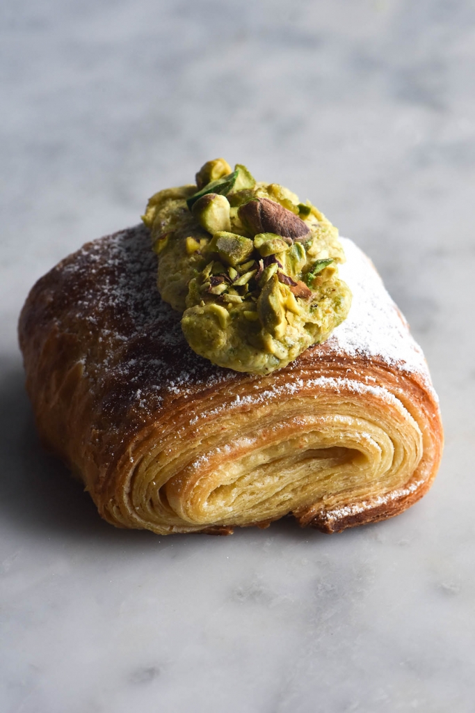 A slightly elevated close up view of a gluten free croissant in the style of a pain au chocolat. The pastry sits atop a white marble table and it's swirled laminated edges face the camera. It is topped with pistachio frangipane, chopped pistachios and a dusting of icing sugar