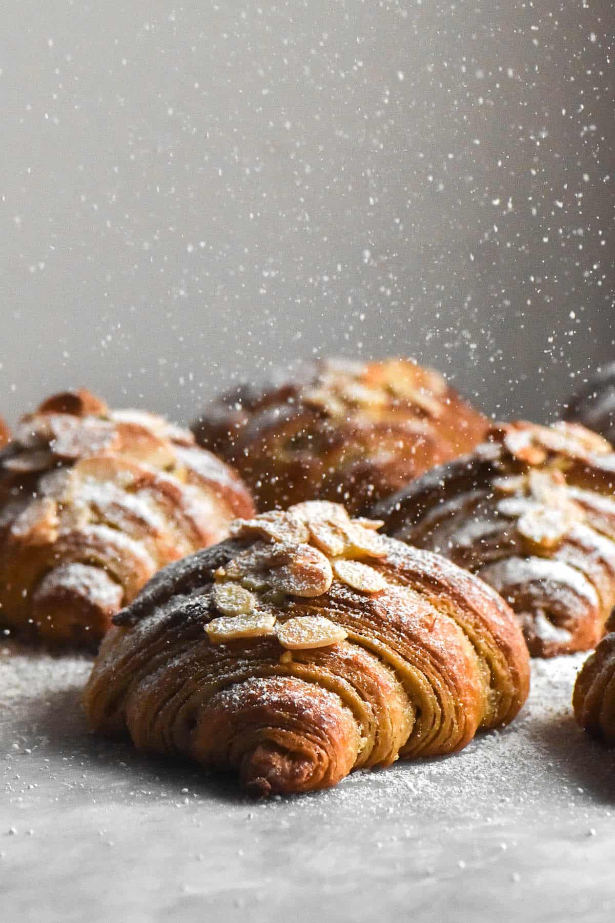 A side on view of a group of gluten free almond croissants being sprinkled with icing sugar. The croissants sit atop a white marble table against a white background, and the sprinkle of icing sugar drops down like flakes of snow. The croissants face the camera to reveal their flaky layers