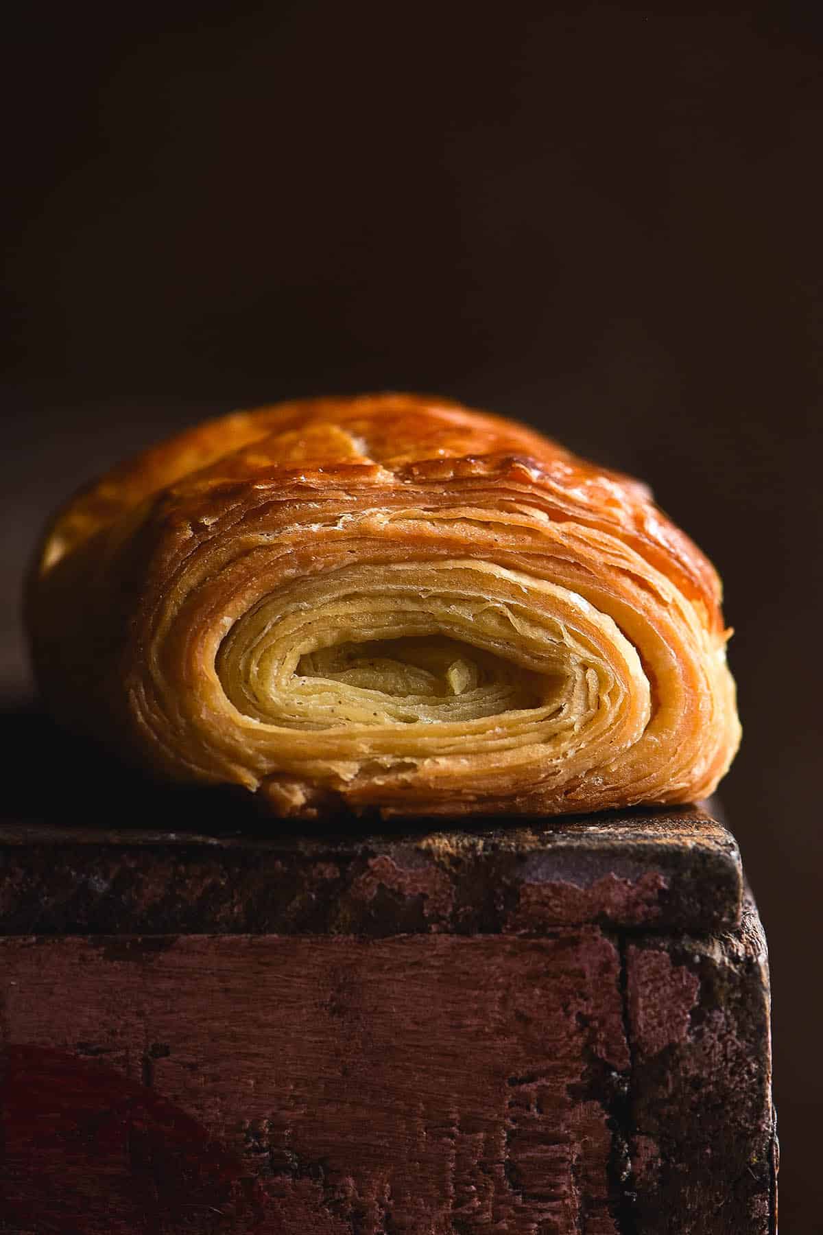 A moody macro side on shot of a gluten free pain au chocolat. The pastry sits on a wooden backdrop with faint pink paint on the side facing the camera. The backdrop is a dark, deep rusty colour which contrasts with the light golden colour of the pastry. The shot focuses on the flaky swirl of layers.