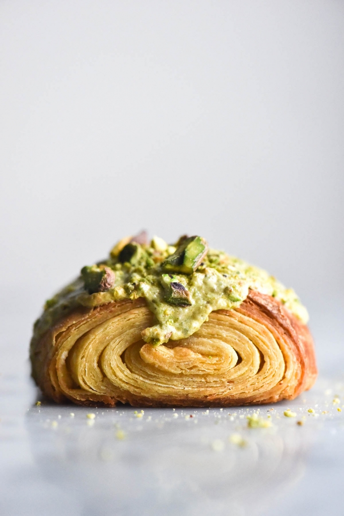 A side on macro photo of a gluten free pain au chocolat topped with pistachio frangipane to make a pistachio croissant. The pastry sits atop a white marble table against a white backdrop. The swirl of the laminated pastry is on display and the light green frangipane drips down the front of the croissant dough. It is topped with extra pistachios, the crumbs of which surround the base of the pastry