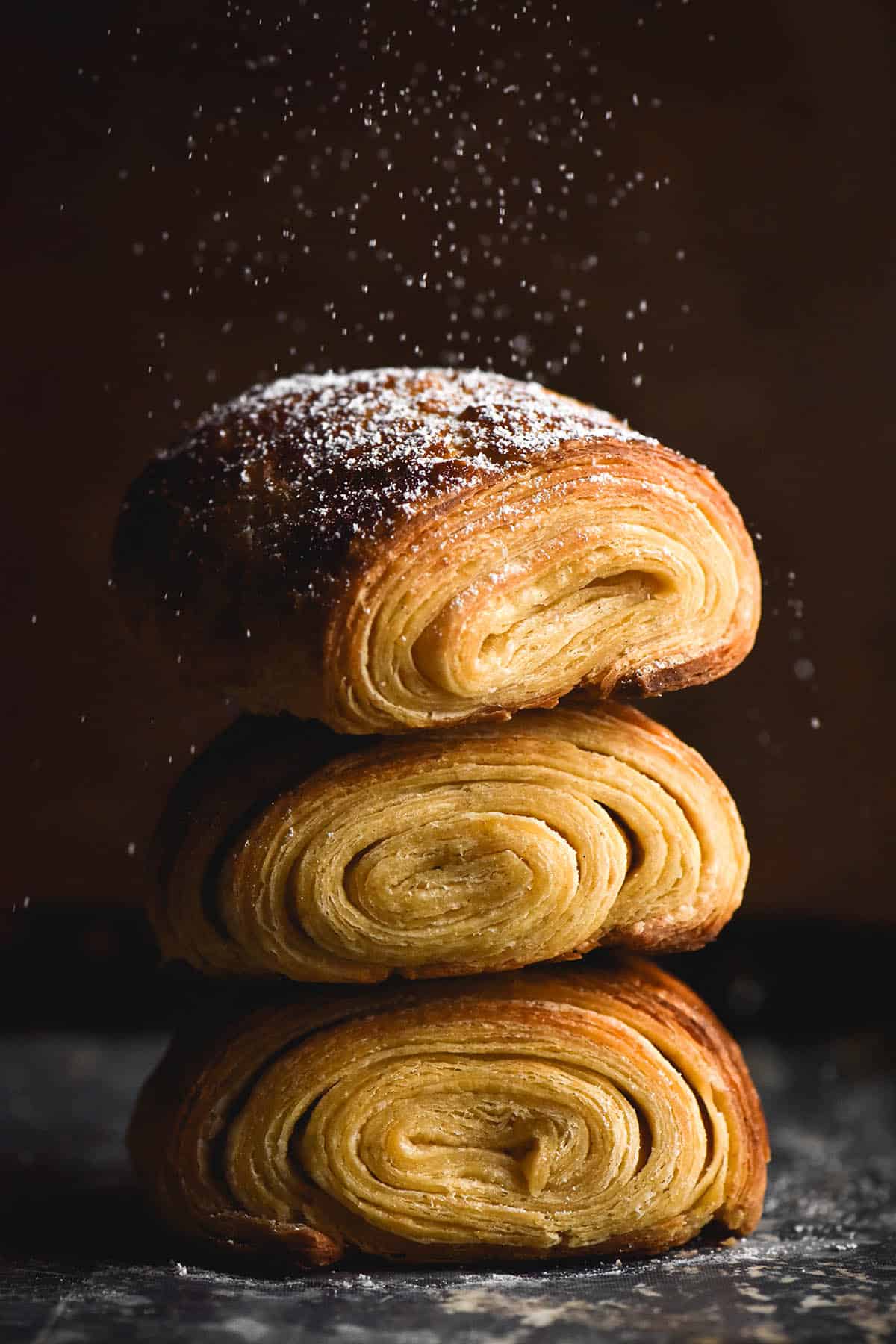 A moody, macro side on view of a stack of three gluten free pains au chocolat. The pastries sit atop a dark coloured baking tray against a dark backdrop. Icing sugar sprinkles from the top of the shot onto the top pastry. The pastries face the camera so that the swirl of lamination is visible