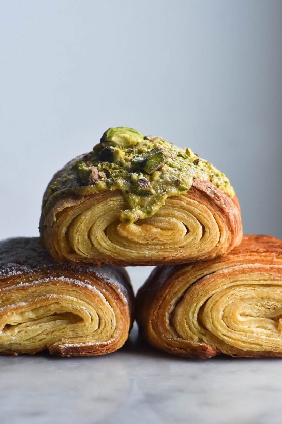 A stack of three gluten free pains au chocolat topped with pistachio frangipane. The pastries sit atop a white marble table against a white backdrop. The pastries sit in a triangle, with the centre pastry resting on each of the two bottom ones.
