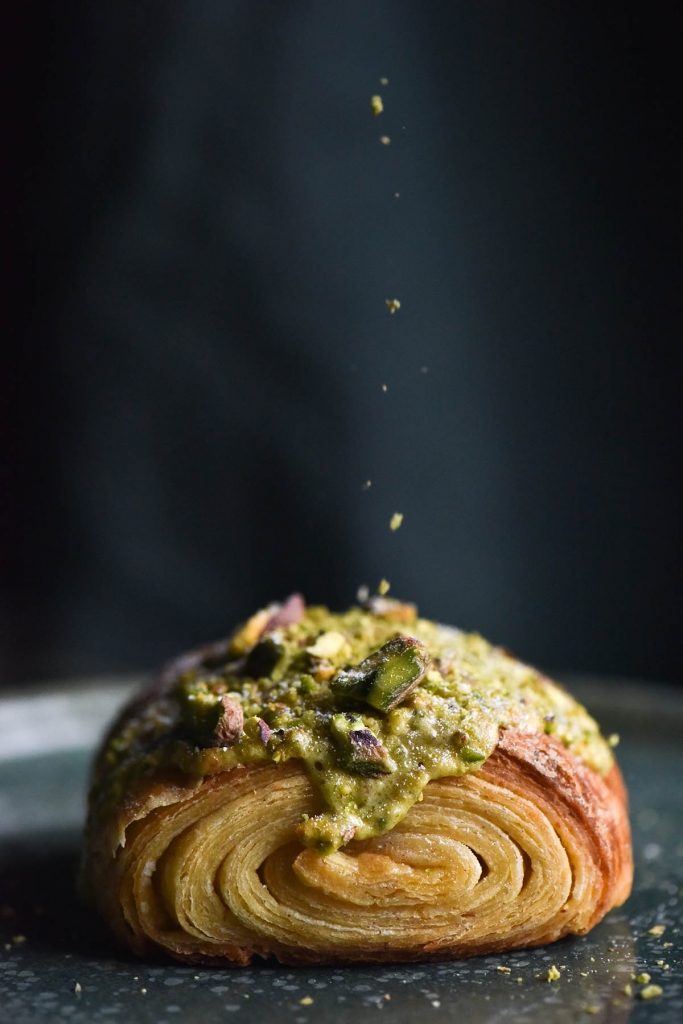 A side on view of a gluten free pain au chocolat topped with pistachio frangipane and extra pistachios. The pastry sits atop a green plate and against an olive green linen backdrop. A thin stream of pistachios are being sprinkled from the top of the shot onto the pain au chocolat