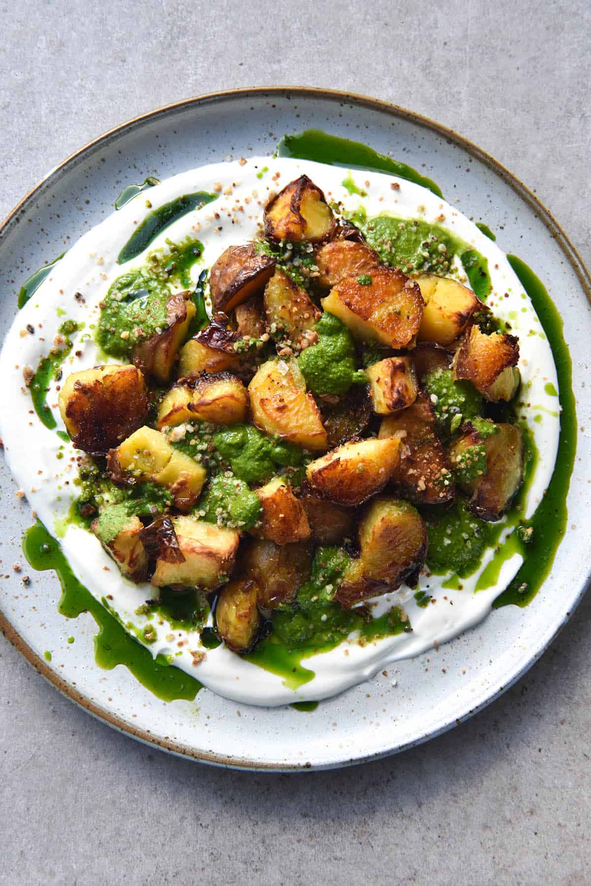 An aerial close up photo of a plate of crispy roast potatoes on a bed of Meredith Goat Cheese dip and topped with FODMAP friendly kale pesto. The green oil from the pesto snakes down the goat cheese spread and onto the plate. The plate is a white ceramic one with a terracotta lip, and it sits atop a light grey stone background