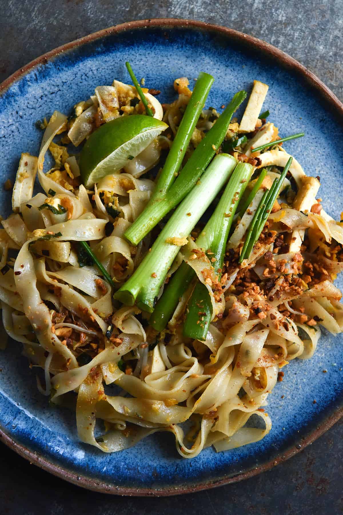 A close up aerial view of a plate of FODMAP friendly Pad Thai. The plate is a vibrant sky blue and it sits atop a darker blue metal backdrop. The Pad Thai is casually arranged and topped with thumb sized pieces of spring onion greens, toasted peanuts and wedges of lime