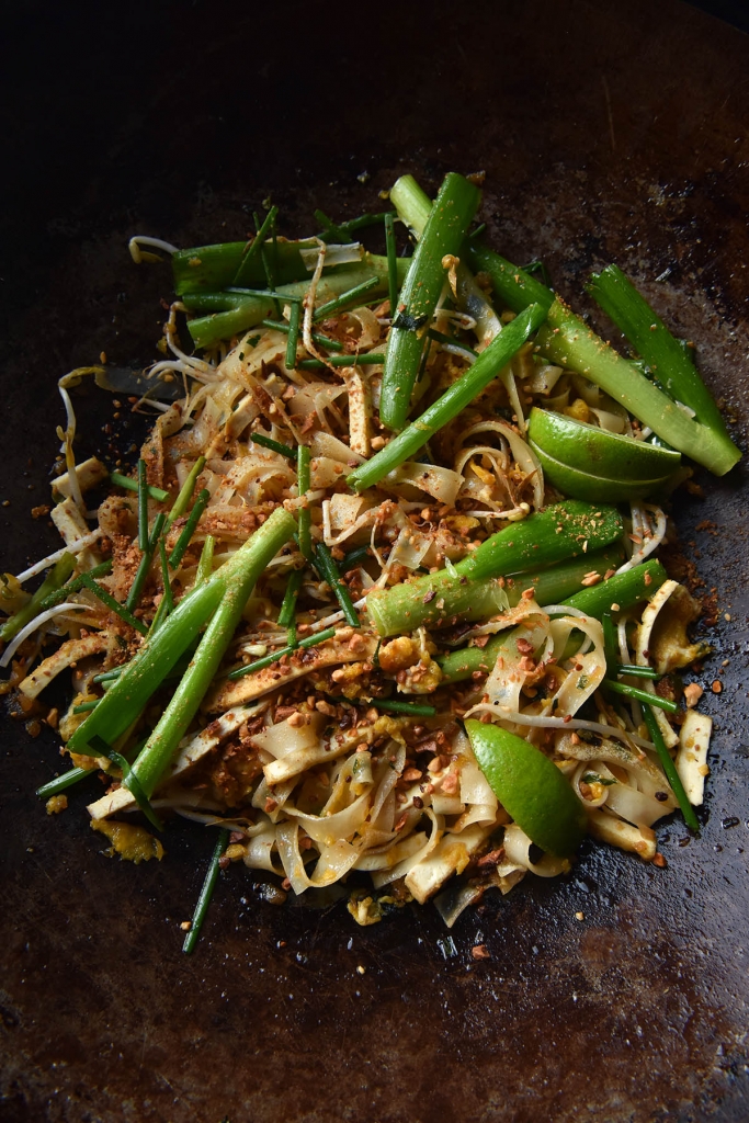 An aerial close up view of FODMAP friendly vegetarian Pad Thai in a rust coloured wok. The Pad Thai is casually arranged in the base of the wok, and is topped with thumb sized spring onion greens, bean sprouts, toasted peanuts and wedges of lime