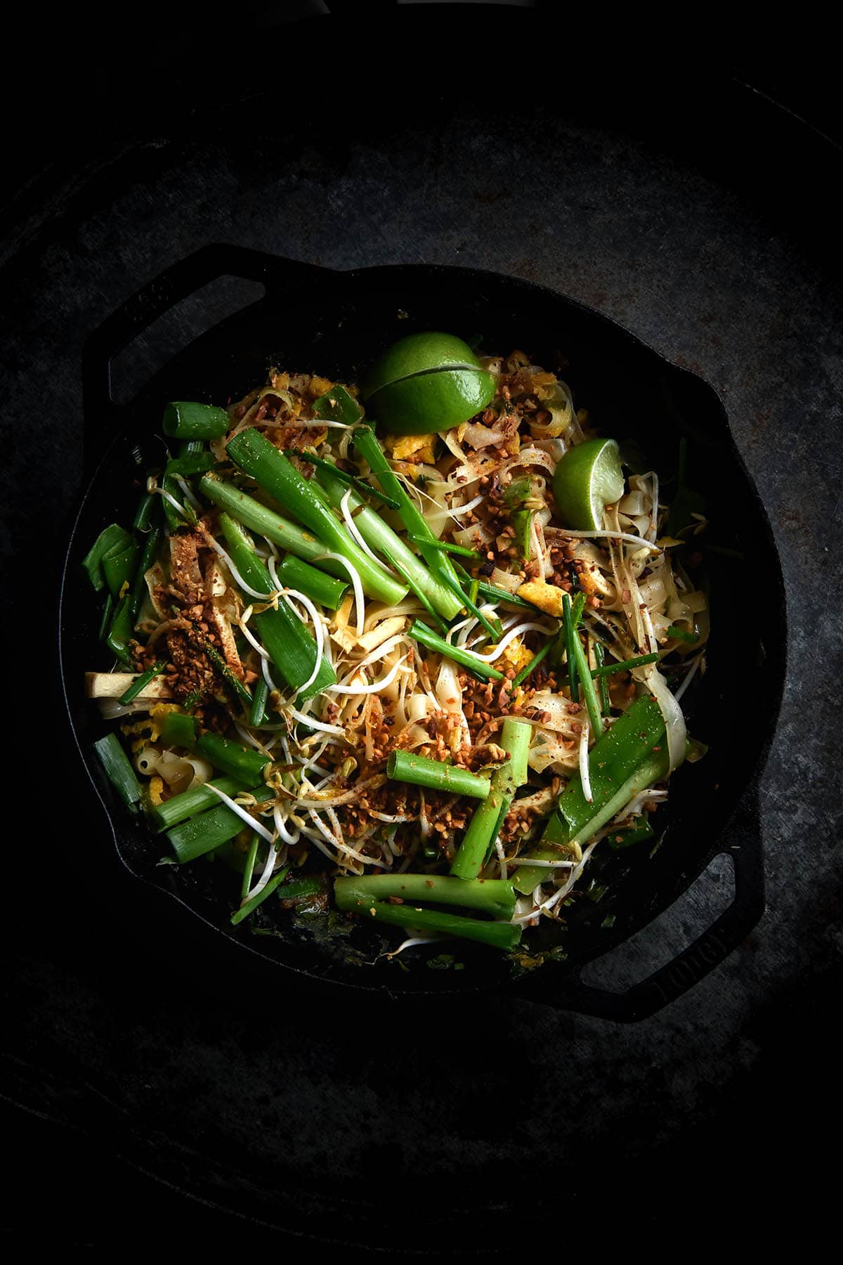 A dark and moody aerial view of a skillet of FODMAP friendly Pad Thai. The Pad Thai sits in the middle of the image and is topped with bean sprouts, spring onion greens, toasted peanuts and lime wedges