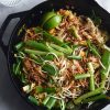 An aerial view of a black skillet filled with FODMAP friendly Pad Thai on a white marble table. A glass of water sits to the top left of the image, catching sunlight. The Pad Thai is casually arranged and topped with spring onion greens, bean sprouts, toasted peanuts and wedges of lime