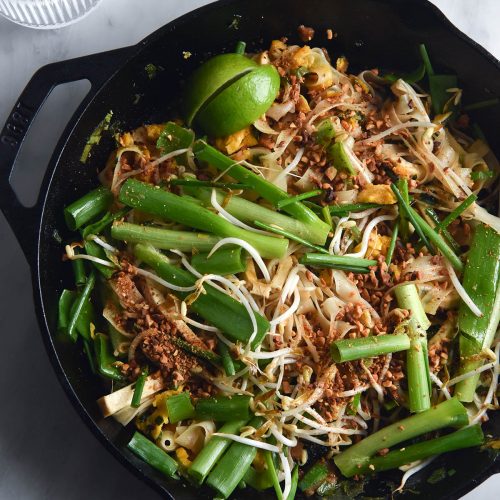 An aerial view of a black skillet filled with FODMAP friendly Pad Thai on a white marble table. A glass of water sits to the top left of the image, catching sunlight. The Pad Thai is casually arranged and topped with spring onion greens, bean sprouts, toasted peanuts and wedges of lime