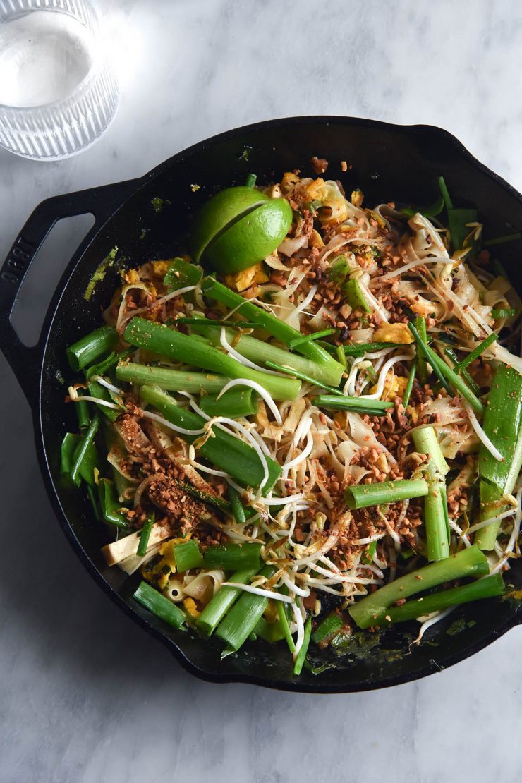 An aerial view of a black skillet filled with FODMAP friendly Pad Thai on a white marble table. A glass of water sits to the top left of the image, catching sunlight. The Pad Thai is casually arranged and topped with spring onion greens, bean sprouts, toasted peanuts and wedges of lime