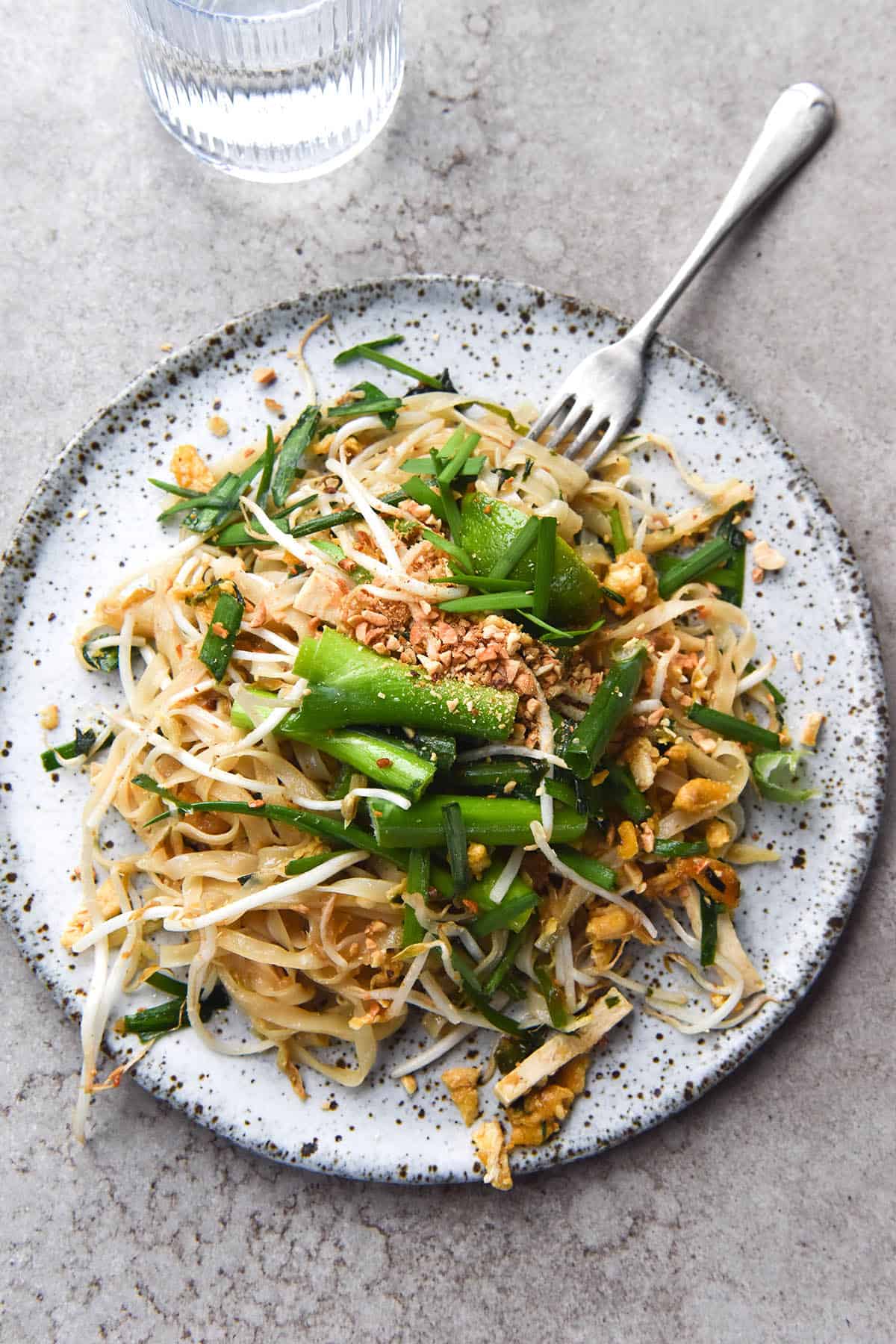 An aerial, light and breezy photo of FODMAP friendly Pad Thai. The Pad Thai sits atop a white speckled ceramic plate on a light grey backdrop. A fork sticks into the top right of the plate, and a glass of water sits to the top left. The Pad Thai is casually arranged and topped with spring onion greens, bean sprouts and toasted peanuts