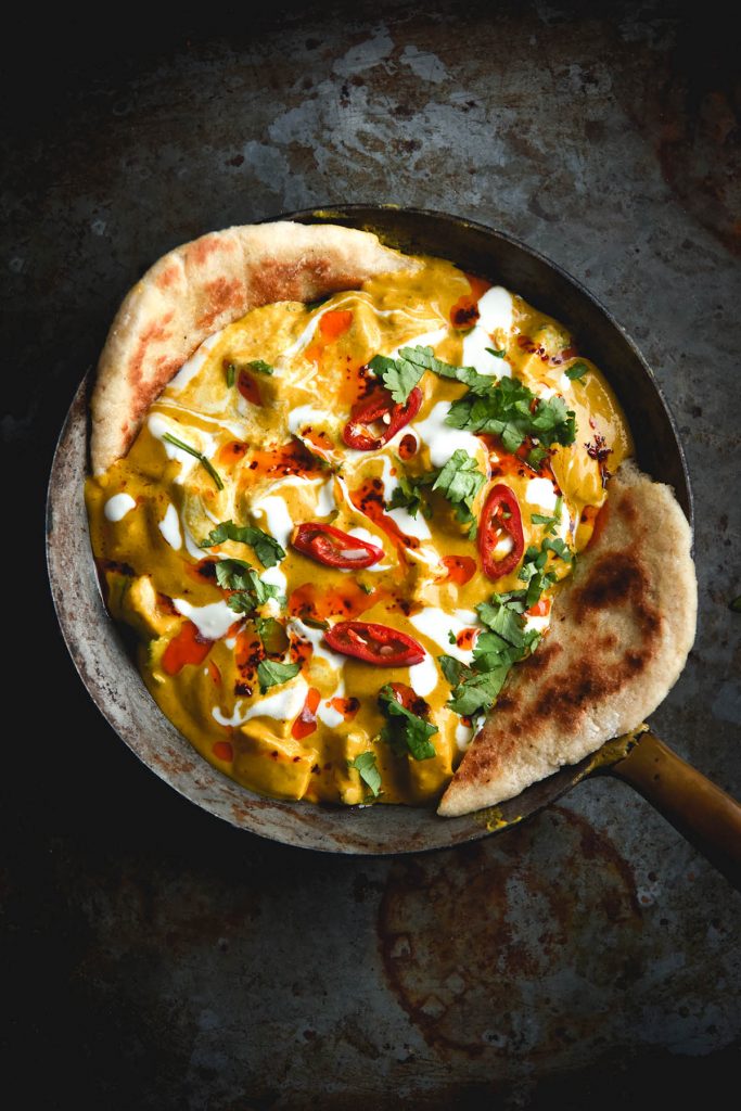 An aerial view of a small skillet filled with FODMAP friendly shahi paneer and adorned with extra cream, sliced chilli and coriander. Two pieces of gluten free yeasted flatbread dip into the curry from the left and right of the image. The skillet sits on a dark metal rusty backdrop