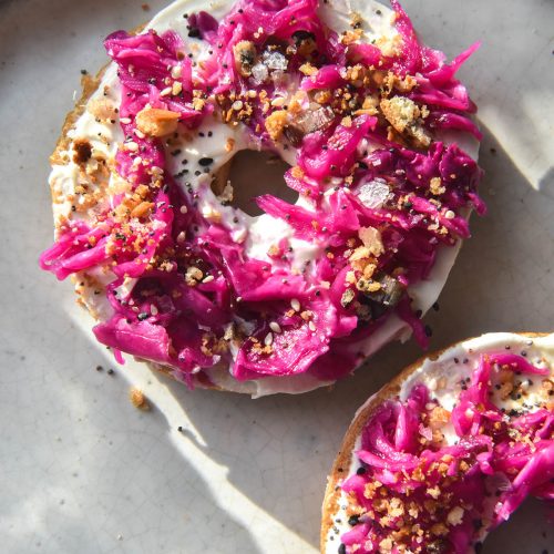 An aerial view of a bagel sliced in half on a white ceramic plate in sunlight. The bagel slices are topped with goats cheese spread, FODMAP friendly 'pickled red onion', FODMAP friendly everything bagel mix and toasted lemon breadcrumbs