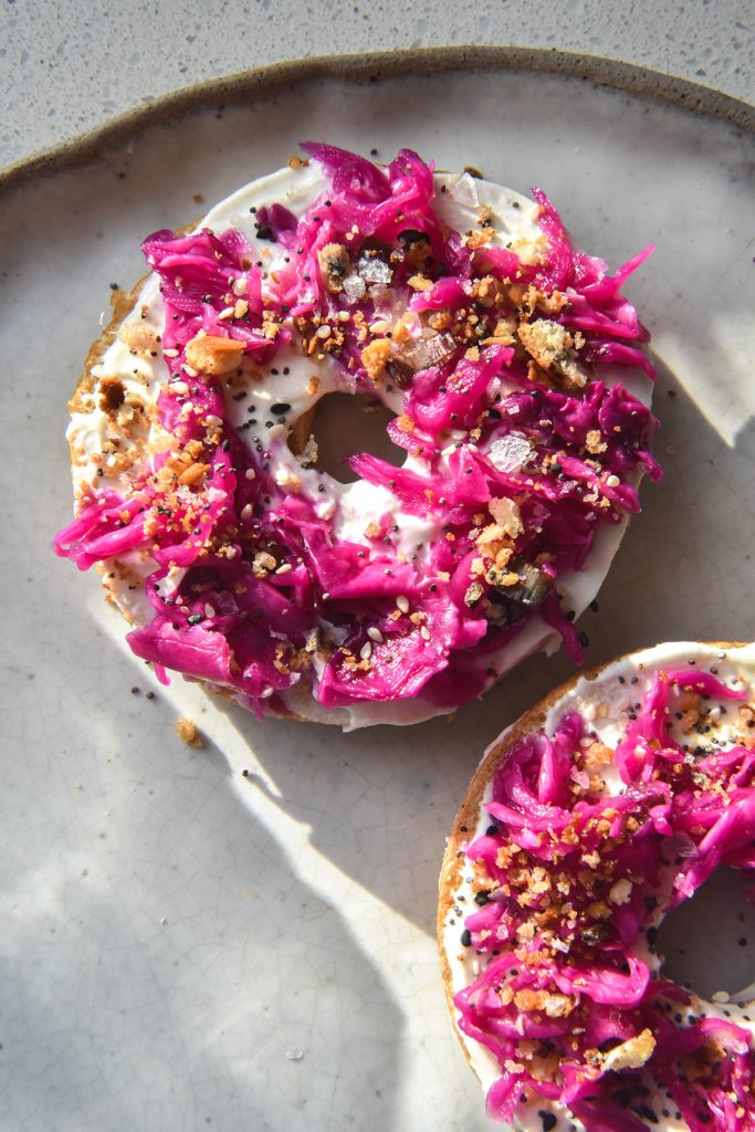 An aerial view of a bagel sliced in half on a white ceramic plate in sunlight. The bagel slices are topped with goats cheese spread, FODMAP friendly 'pickled red onion', FODMAP friendly everything bagel mix and toasted lemon breadcrumbs