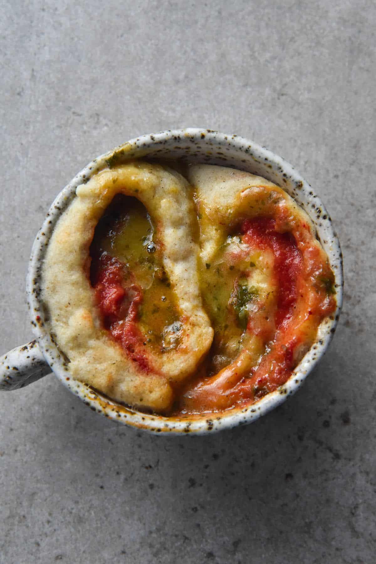 An aerial close up view of a pizza flavoured mug cake. The background is a light grey stone colour and atop that sits the white speckled mug. Inside the mug is two swirls of a gluten free savoury scroll which has been cooked in the microwave. The scrolls are filled with pizza sauce, pesto and melty cheese, and the ingredients have mingled together to form different hues during the cooking process
