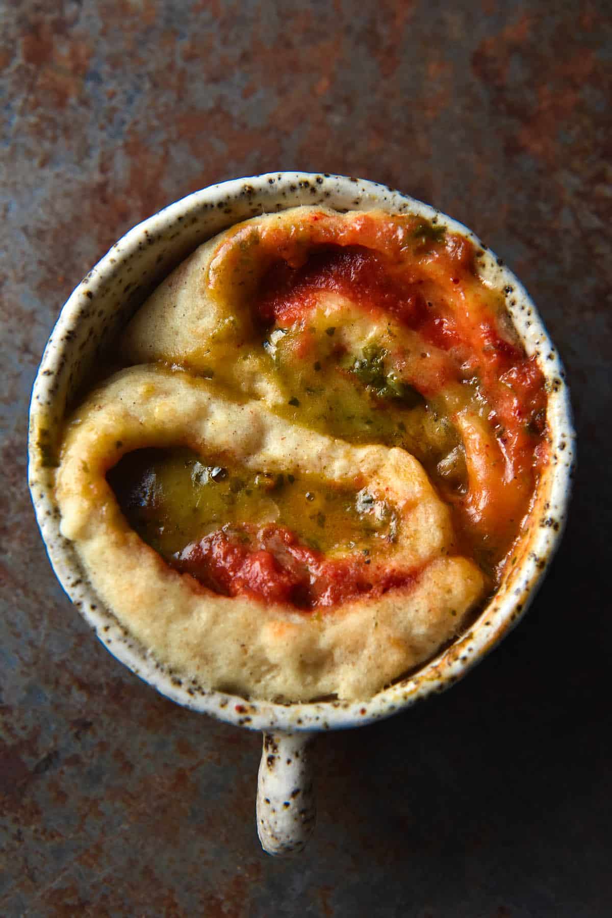 An aerial close up view of a pizza flavoured mug cake. The background is a rusted denim blue colour and atop that sits the white speckled mug. Inside the mug is two swirls of a gluten free savoury scroll which has been cooked in the microwave. The scrolls are filled with pizza sauce, pesto and melty cheese, and the ingredients have mingled together to form different hues during the cooking process