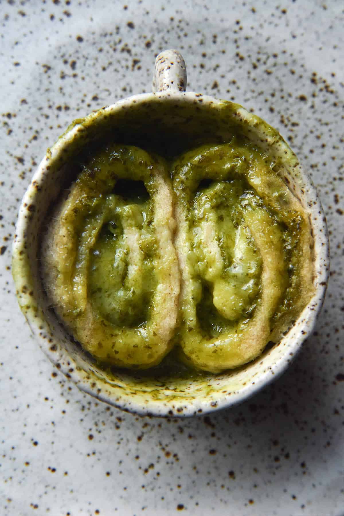 An aerial close up photo of a pesto scroll mug cake in a white speckled ceramic mug on a white speckled ceramic plate. The scroll has been sliced into two before baking, leaving two scrolls snuggled into eachother and facing upwards in the mug. The pesto and cheese have melted together to create an oozy, gooey and delicious swirled result
