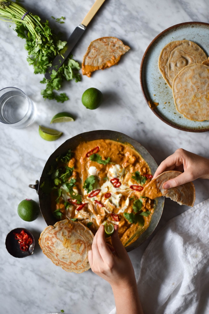 An aerial view of a dish of FODMAP friendly shahi paneer on a white marble table, surrounded by plates, glasses, extra coriander and limes. Two hands dip a piece of flatbread into the shahi paneer from the bottom left hand side of the image.