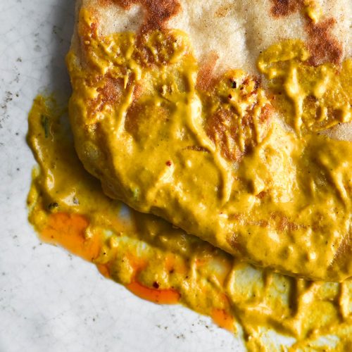 An aeria close up view of a gluten free yeasted flatbread that has been dipped in the gravy of a FODMAP friendly shahi paneer. It sits atop a white ceramic plate and the gravy is smeared across the bottom portion of the plate