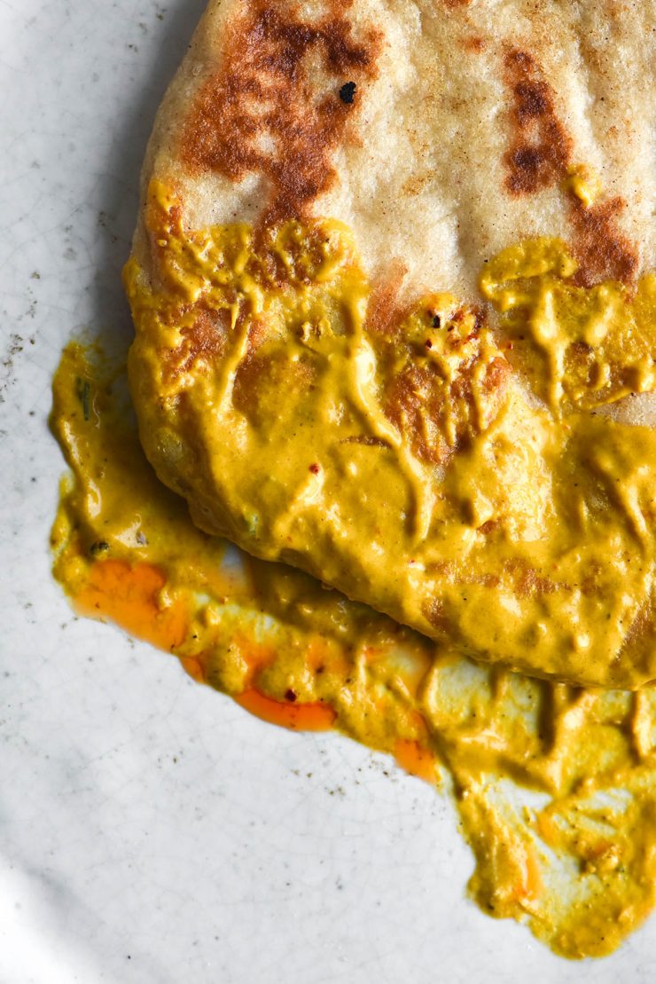 An aeria close up view of a gluten free yeasted flatbread that has been dipped in the gravy of a FODMAP friendly shahi paneer. It sits atop a white ceramic plate and the gravy is smeared across the bottom portion of the plate