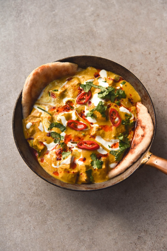 An aerial image of a small skillet filled with FODMAP friendly shahi paneer. The shahi paneer has two pieces of flatbread sticking out of each side. It is topped with extra cream, coriander and chopped chilli. The skillet sits atop a light grey stone backdrop in the centre of the image