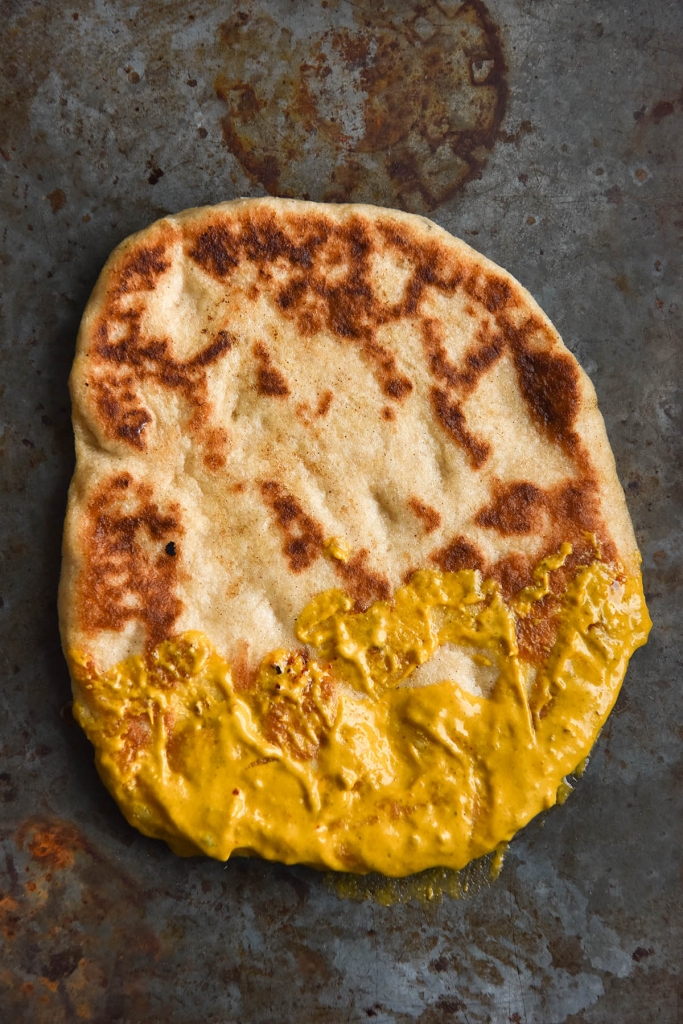 An aerial view of a gluten free flatbread half dipped on curry gravy. The flatbread is on a rustic grey and rust coloured backdrop and the bottom third of the flatbread is covered in rich, bright orange paneer shahi gravy