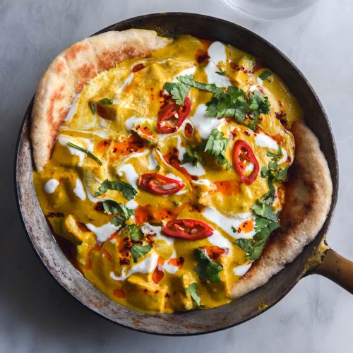 An aerial view of a small skillet filled with FODMAP friendly shahi paneer. The shahi paneer is topped with extra cream, chopped coriander and sliced chillies. Two pieces of flabread stick out of the curry on the left and right sides of the skillet. The skillet sits atop a white marble table, and some sunlit water glasses sit in the top left of the image