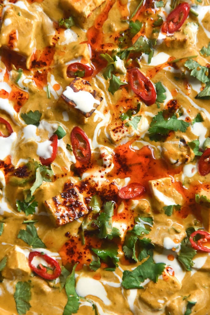 An aerial macro view of the surface of a skillet filled with FODMAP friendly shahi paneer. The dish is dotted with sliced red chilli, coriander, and drizzled with extra cream and a bit of chilli oil