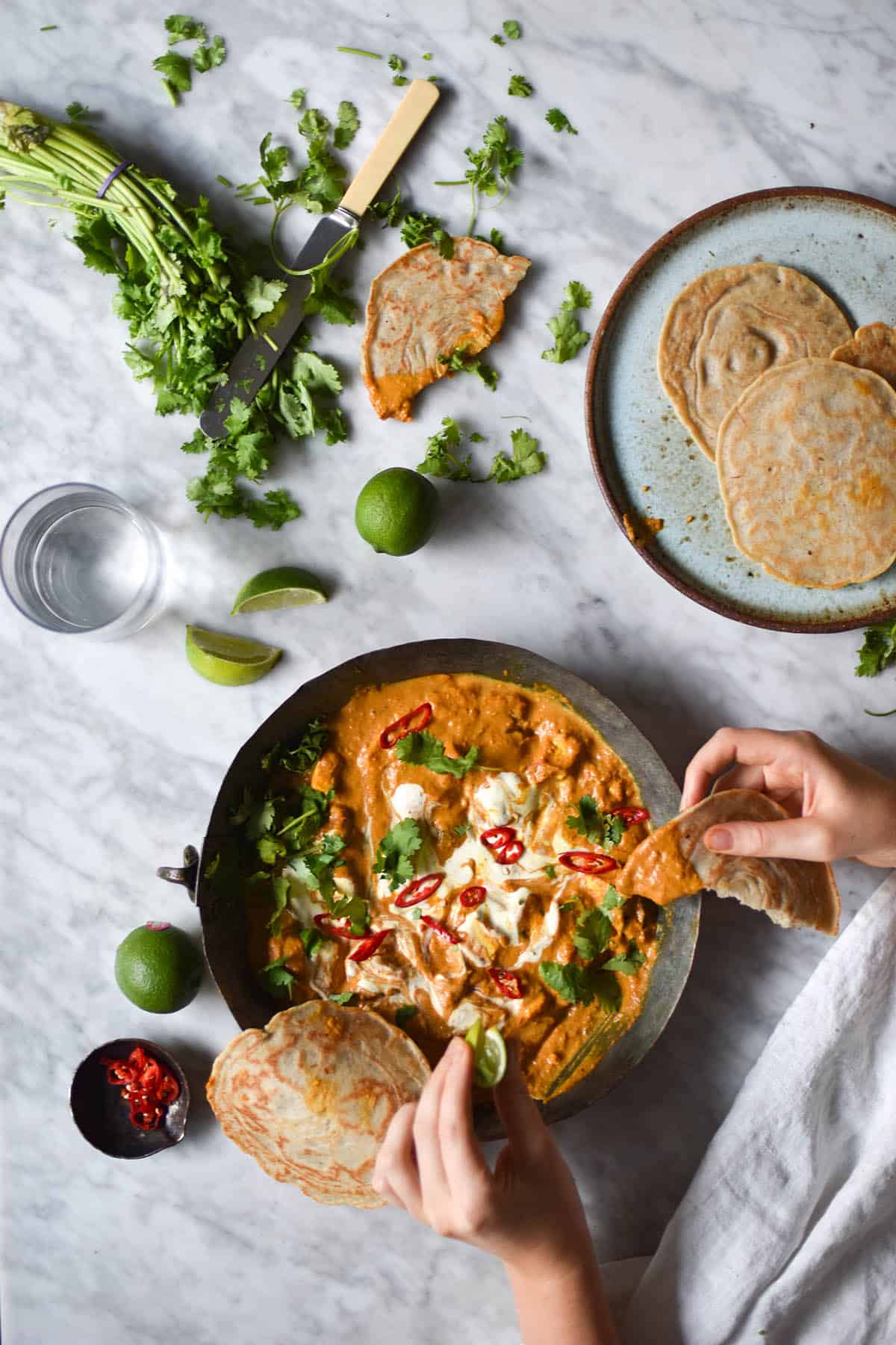 An aerial view of a marble table topped with a metal dish of FODMAP friendly shahi paneer, surrounded by casually strewn limes, chilli, coriander and gluten free yeasted flatbread. Two hands extend from the bottom left of the image to tear a flatbread and dip it into the curry, which is topped with coriander, chillies and a swirl of cream
