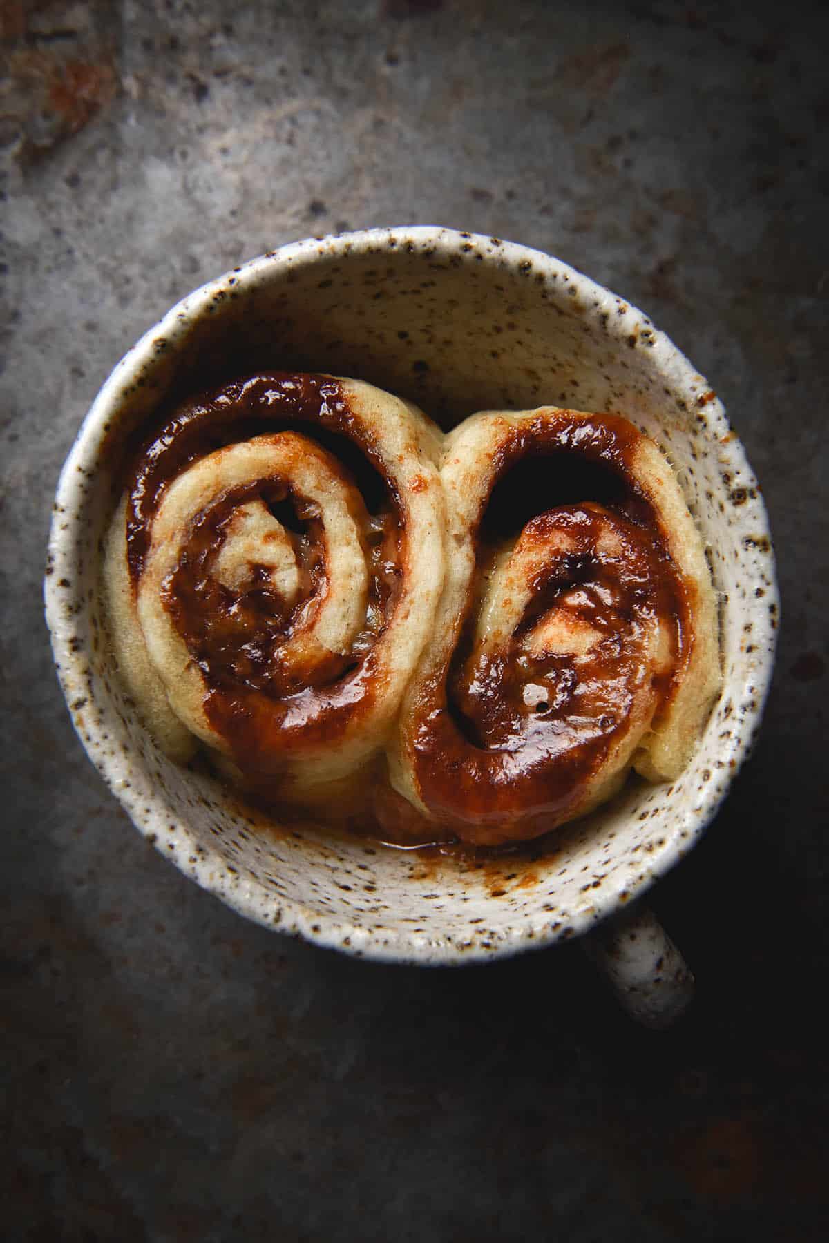 An aerial view of a gluten free Vegemite scroll made in a mug. The mug is a white speckled ceramic one that sits atop a dark grey rusty backdrop. The single serve scroll was sliced into two before baking, leaving two oozy, melty, cheesy scrolls snuggled up against each other in the mug