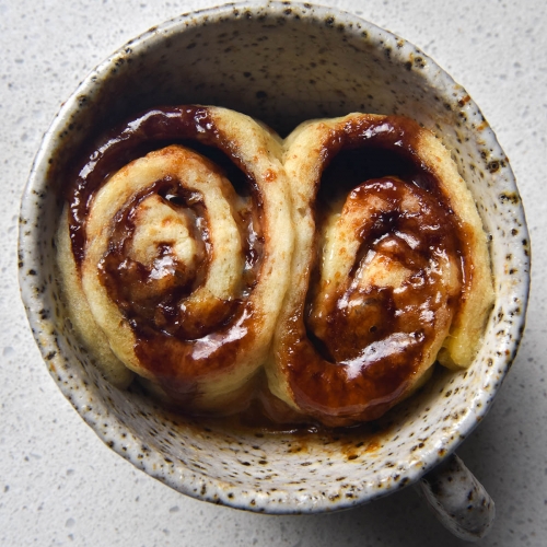 An aerial view of a gluten free Vegemite scroll made in a mug. The mug is a white speckled ceramic one that sits atop a white kitchen counter. The single serve scroll was sliced into two before baking, leaving two oozy, melty, cheesy scrolls snuggled up against eachother in the mug