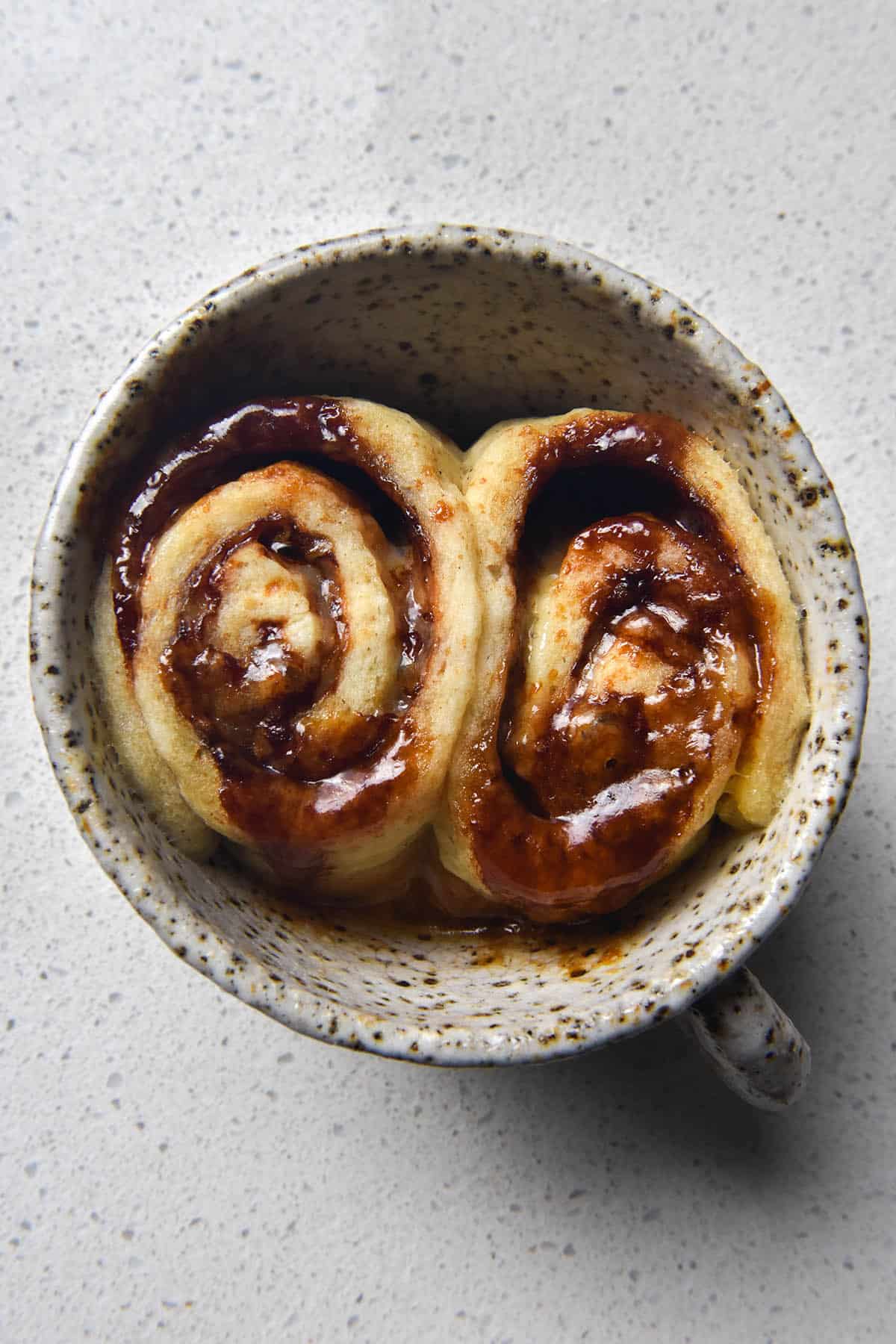 An aerial view of a gluten free Vegemite scroll made in a mug. The mug is a white speckled ceramic one that sits atop a white kitchen counter. The single serve scroll was sliced into two before baking, leaving two oozy, melty, cheesy scrolls snuggled up against eachother in the mug