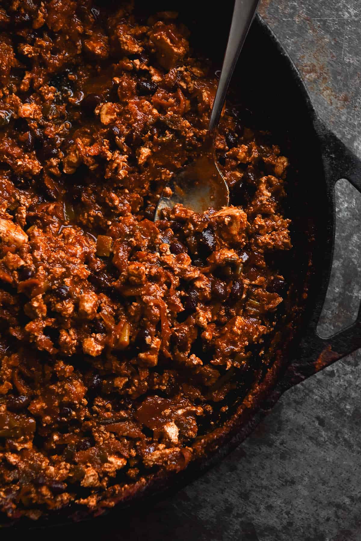 An aerial close up view of a skillet filled with FODMAP friendly vegetarian chilli sin carne. The skillet sits on a dark grey metal backdrop for a moody image. A spoon rests in the chilli in the top righthand corner of the image and extends out to the top left