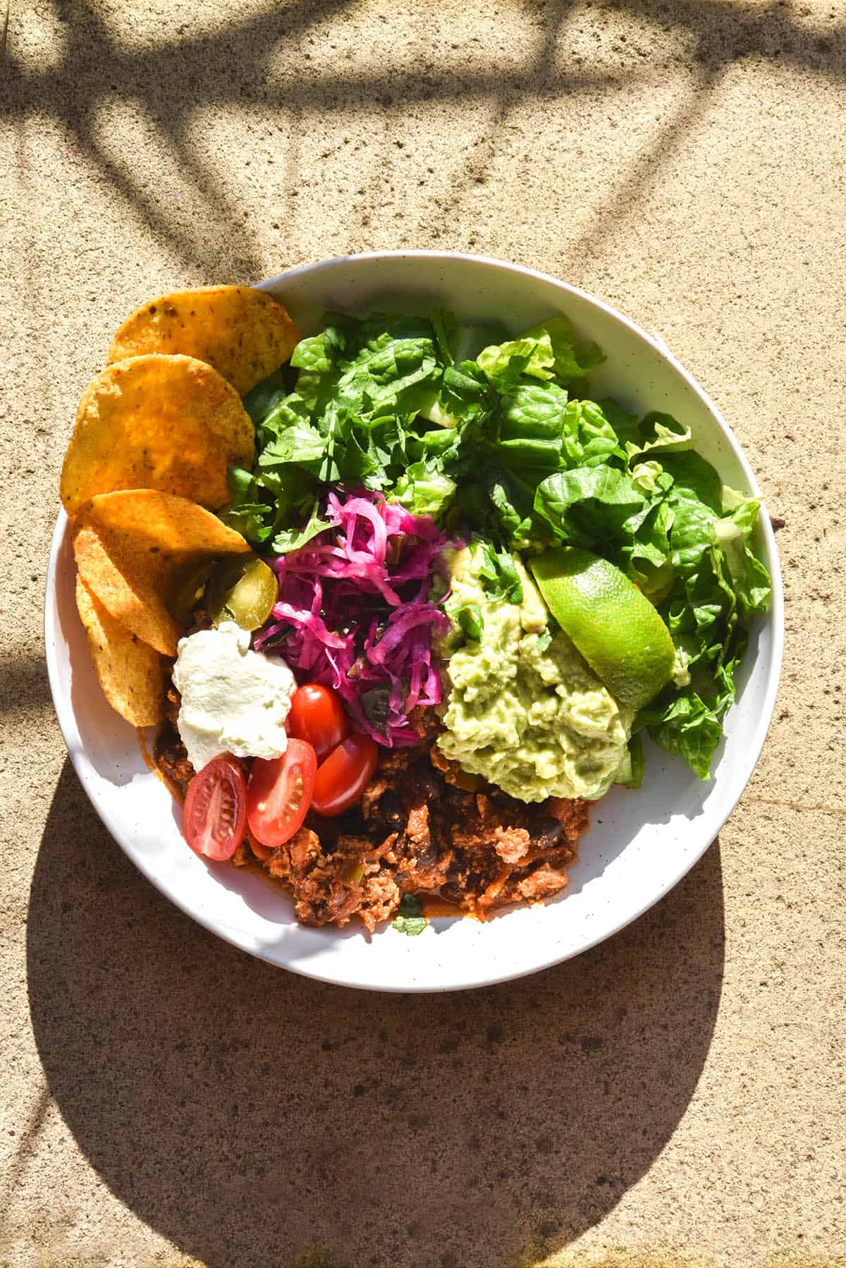 An aerial brightly lit view of a plate of FODMAP friendly vegetarian chilli served with salad, guacamole, a wedge of lime, some fodmap friendly 'pickled red onion', corn chips, sour cream and tomatoes. It sits atop terracotta tiles in bright sunlight, with the shadow of some palm leaves in the background of the image
